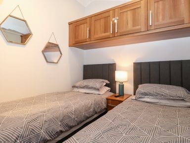 A bedroom with two single beds, a bedside table with a lamp, wooden overhead cabinets, and two hexagonal mirrors on the wall at Squirrels Nook Waterside Wood 19 White Cross Bay near Windermere
