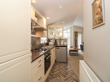 A narrow kitchen with cream cabinets and appliances leading to a living area with a coffee table and armchair at Squirrels Nook Waterside Wood 19 White Cross Bay near Windermere