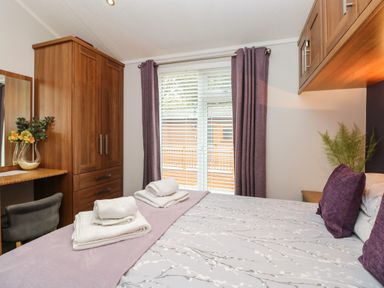 A bedroom with a bed, folded towels, wooden wardrobe, dressing table with a chair, window with blinds and purple curtains at Squirrels Nook Waterside Wood 19 White Cross Bay near Windermere