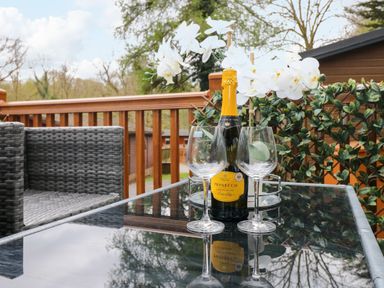 A glass table with a bottle of prosecco and two empty wine glasses on a patio with gray wicker chairs and a wooden railing at Squirrels Nook Waterside Wood 19 White Cross Bay near Windermere