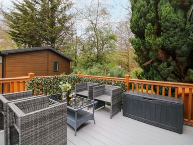 An outdoor deck with wicker chairs glass table and a storage box surrounded by trees at Squirrels Nook in Waterside Wood 19 White Cross Bay near Windermere