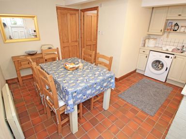 A dining area with a table covered by a blue tablecloth and wooden chairs beside a kitchen with white cabinets and a washing machine at Hafan Cottage in Beddgelert