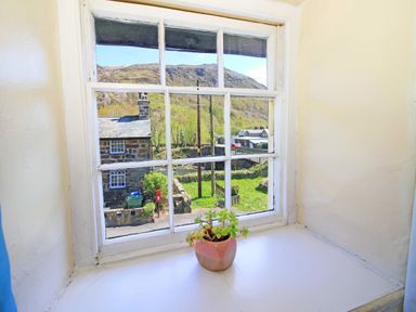 A window with white frame and a potted plant on the sill overlooking a stone house and a green hillside at Hafan Cottage in Beddgelert