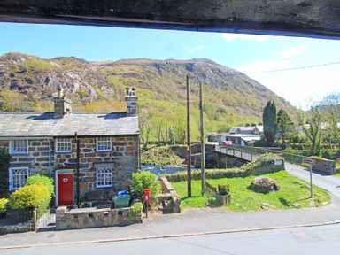A stone cottage with a red door beside a river and a bridge with hills in the background at Hafan Cottage in Beddgelert