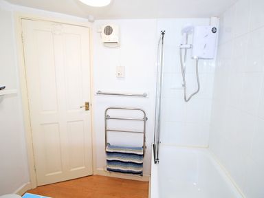 A bathroom with a bathtub and shower unit a towel rack with a striped towel a door and a wall-mounted ventilation fan at Hafan Cottage in Beddgelert