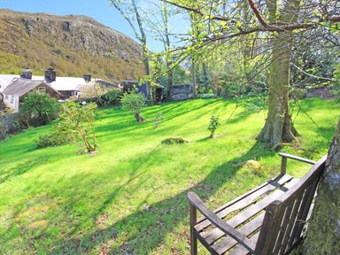 An outdoor garden with grass shrubs and trees a wooden bench and houses near a mountain at Hafan Cottage in Beddgelert