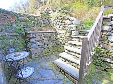An outdoor stone patio with mosaic table and chairs and stone steps with wooden handrail at Hafan Cottage in Beddgelert