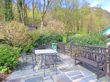 An outdoor patio with metal table and chairs and wooden benches surrounded by trees and bushes at Hafan Cottage in Beddgelert