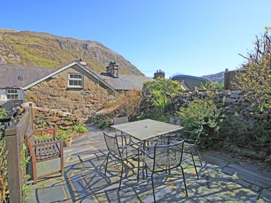 An outdoor patio with metal table and chairs a wooden bench stone wall and hills in the background at Hafan Cottage in Beddgelert