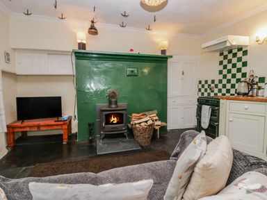 A living room area with a wood burning stove and firewood basket next to a kitchen area with stove and kettle at Gardeners Cottage in Millom Cumbria