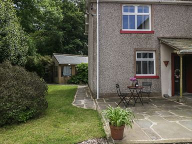 An outdoor patio area with two chairs and a table with flowers and a bottle next to a house and a small shed in the garden at Gardeners Cottage in Millom Cumbria