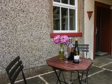 A small outdoor table with two chairs beside a pebble wall with a window a vase with flowers and two bottles on the table at Gardeners Cottage in Millom Cumbria
