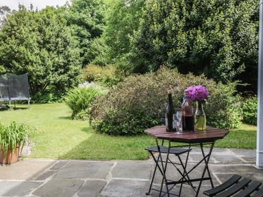 A garden with green bushes and trees a small wooden table with bottles glasses and flowers on a stone patio