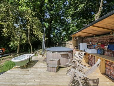 An outdoor wooden deck with a clawfoot bathtub a round hot tub two wooden chairs and a wooden counter with a sink at Gardeners Cottage in Millom Cumbria