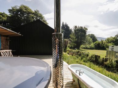 An outdoor area with a round hot tub cover a metal chimney pipe a clawfoot bathtub filled with water on a wooden deck and green trees at Gardeners Cottage Millom Cumbria