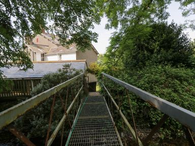 A metal footbridge over a stream leading to a house surrounded by trees and bushes at Gardeners Cottage in Millom Cumbria