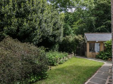 A garden with bushes and trees a grass lawn a wooden shed and a stone pathway at Gardeners Cottage in Millom Cumbria