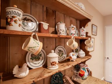 A wooden kitchen shelf with ceramic jars, hanging mugs, decorative plates, and a glass pitcher at 3 Albion Cottages in Walberswick