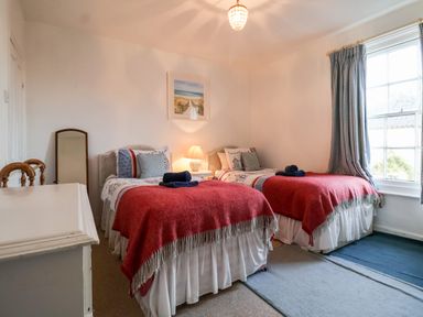 A bedroom with two single beds covered with red blankets a bedside table with a lamp a window with check curtains and a dresser at 3 Albion Cottages in Walberswick