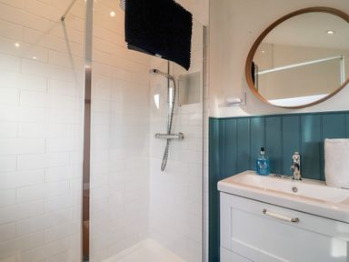 A bathroom with a shower area featuring white tiled walls a sink with a faucet a round mirror and a bottle of blue soap at 3 Albion Cottages in Walberswick