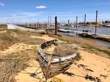 An old wooden boat on sandy ground near a dock with boats and buildings in the background at 3 Albion Cottages in Walberswick