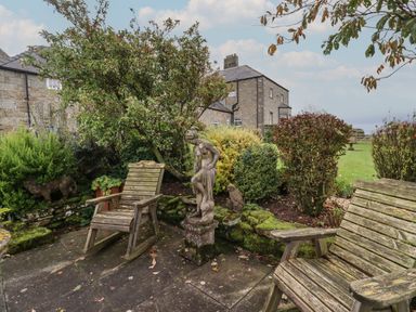 A garden patio with wooden rocking chairs and stone statues surrounded by bushes at The Barn in Middleton near Ponteland