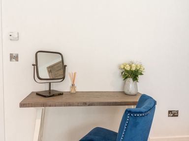 A wooden table with a mirror a reed diffuser and a vase with white roses and a blue chair at 31 Cliff Edge in Newquay