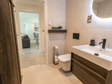 A bathroom with a toilet paper holder a wall-mounted toilet a sink with a black faucet and a round illuminated mirror at 31 Cliff Edge in Newquay