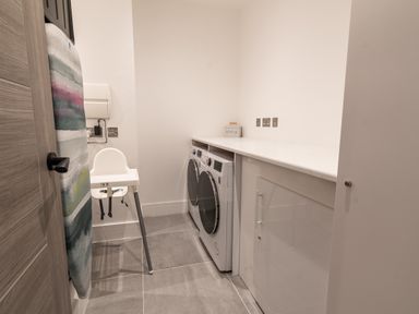 A laundry room with a washing machine, dryer, cabinet, ironing board, and a high chair at 31 Cliff Edge in Newquay