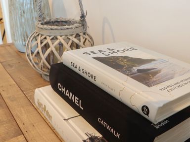 A stack of three books on a wooden surface next to a woven lantern and a ceramic vase