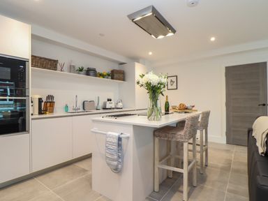 A modern kitchen with white cabinets a kitchen island with flowers and two woven bar stools at 31 Cliff Edge in Newquay