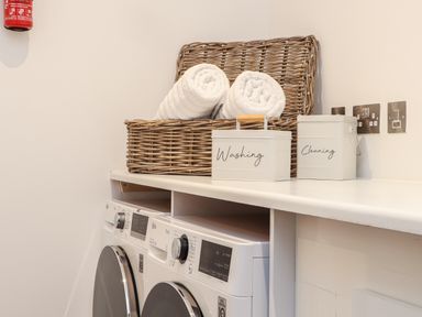A laundry area with a washing machine and dryer below a counter holding a wicker basket with towels and two containers labeled washing and cleaning at 31 Cliff Edge in Newquay