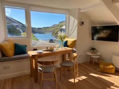 A dining area with a wooden table and chairs by a large window showing a water and hill view at Harbour Light in Boscastle