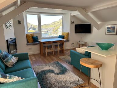 A living room with teal sofas a wooden dining table and chairs a television and window seat with cushions at Harbour Light in Boscastle