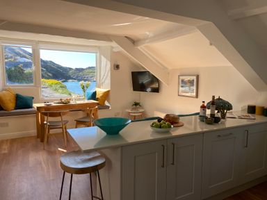 A kitchen with a white island counter and a dining table by a window with a view of water and hills at Harbour Light in Boscastle