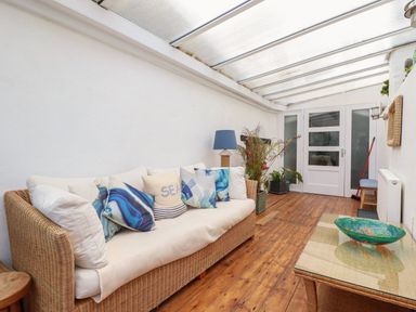 A sunroom with a wicker sofa with cushions a glass coffee table with a decorative bowl plants and a wooden floor at Harbour Light in Boscastle