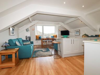 A living area with a teal sofa, wooden side table, a dining table by a window with cushions, a kitchen island with a stool and a TV on the counter at Harbour Light in Boscastle