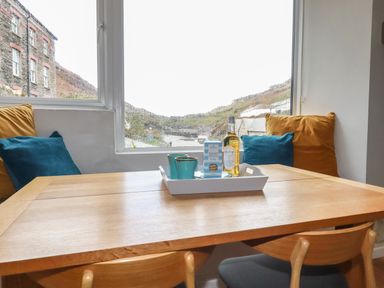 A wooden dining table with chairs and cushions by a window with a tray containing cups a bottle and a box at Harbour Light in Boscastle