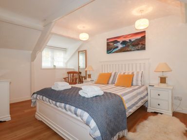 A bedroom with a double bed with striped bedding and yellow pillows two bedside tables with lamps a wooden chair a full-length mirror and a landscape painting on the wall at Harbour Light in Boscastle