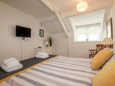 A bedroom with a bed striped bedspread and yellow pillows a dresser with a mirror and plant a chair a table lamp and a wall mounted tv at Harbour Light in Boscastle