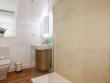 A bathroom with a toilet, sink, towel radiator, shower area with glass partition, and a window with blinds at Harbour Light in Boscastle