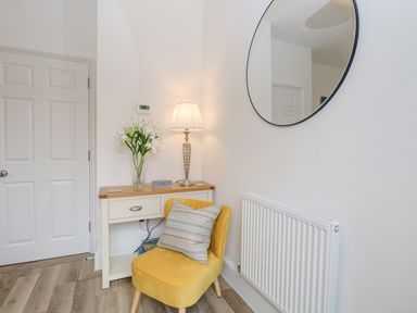 A corner with a white door a small wooden desk with a lamp and flowers a yellow chair with a cushion and a round mirror on the wall at The Hollies in Norwich