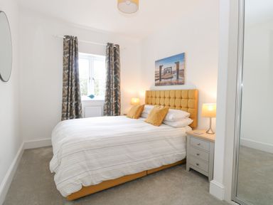 A bedroom with a double bed with yellow headboard and pillows bedside tables with lamps curtains on window and a painting on the wall at The Hollies in Norwich