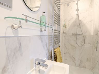 A bathroom with a sink faucet a glass shelf with soap a towel rail with a towel and a shower enclosure with a showerhead and marble wall tiles at The Hollies in Norwich