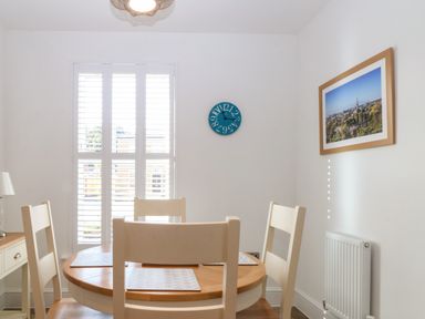 A dining area with a round wooden table and four white chairs by a window with shutters a blue clock on the wall and framed picture at The Hollies in Norwich