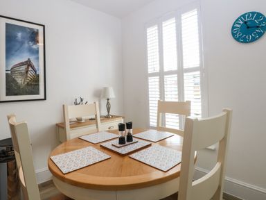 A dining area with a round wooden table set with four placemats and salt and pepper shakers in front of a window with white shutters at The Hollies in Norwich