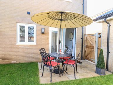 A small patio with a round table and four chairs with red cushions under a large umbrella next to a brick house and a wooden gate at The Hollies in Norwich