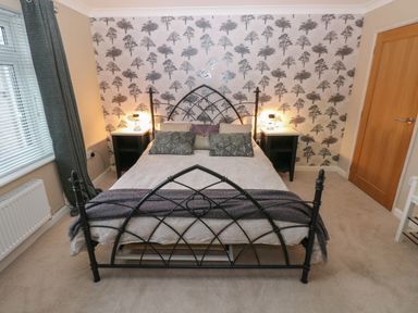 A bedroom with a metal bed frame two bedside tables with lamps tree patterned wallpaper and a window with curtains at Garden View in Pembroke