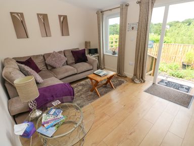 A living room with a beige sofa and purple cushions a small wooden coffee table a glass side table with books and a lamp wooden floor and sliding glass doors leading outside at Garden View in Pembroke