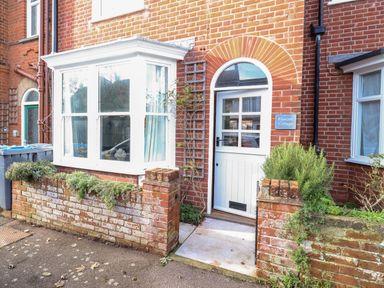 The exterior entrance of Fawcett Cottage with a white door, bay window, brick wall, and plants at Aldeburgh